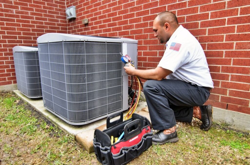 Technician kneeling down, repairing an HVAC unit outside a brick home. Tool bag next to him.