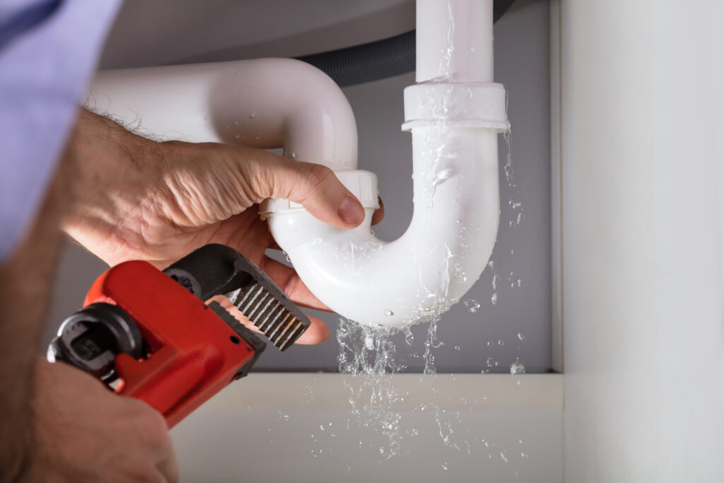 Close-up of plumber's hands fixing white sink pipe with adjustable wrench while pipe is leaking water.